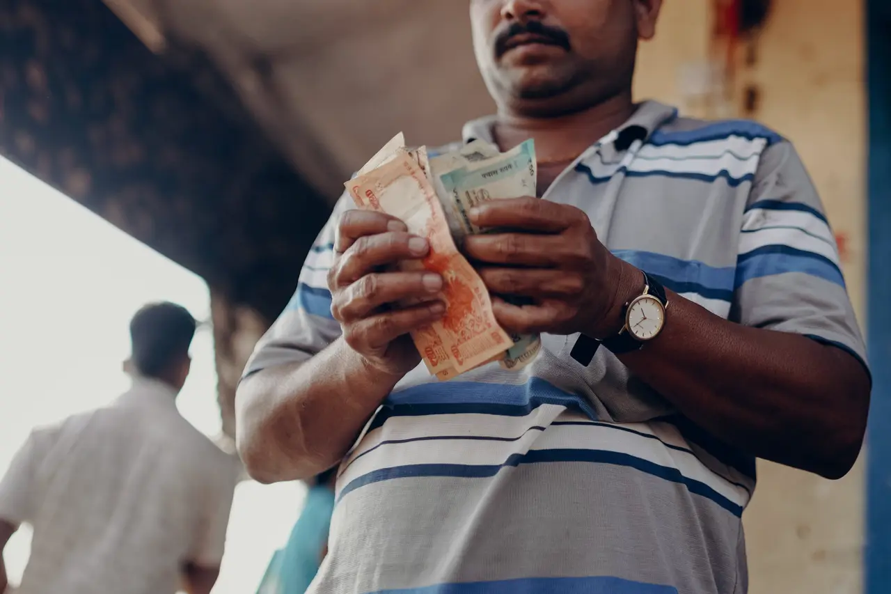 Close-up of a man in a striped polo shirt counting a fold of rupee notes at a market, his face out of the frame.