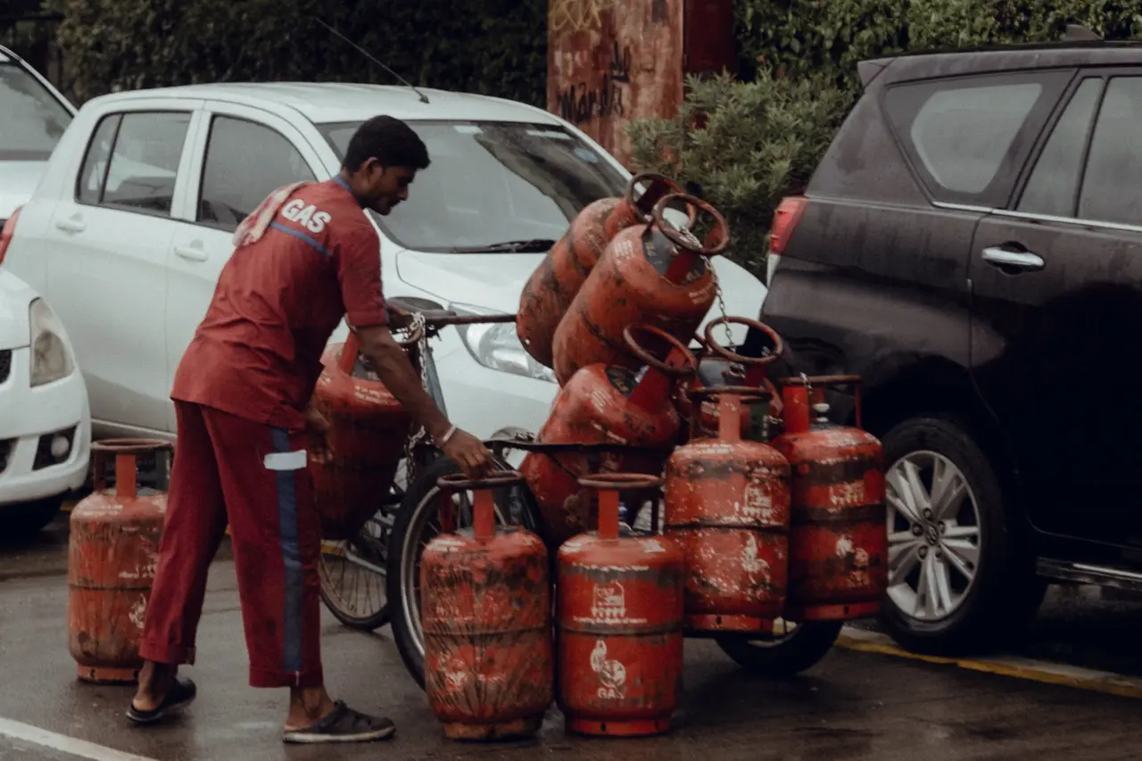 A delivery worker in a red uniform steadies a bicycle loaded with gas cylinders on a wet city street, between parked cars.