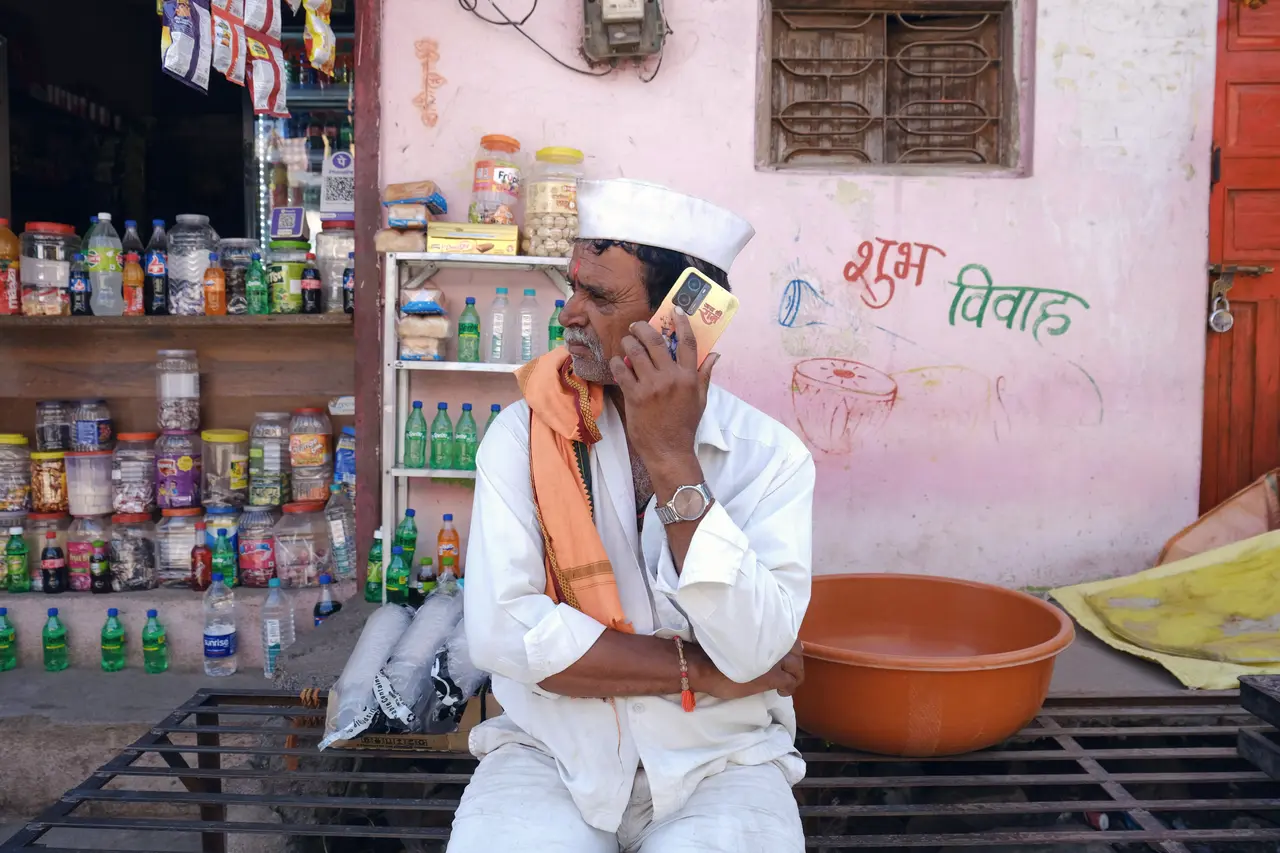 A shopkeeper in a white Gandhi cap and an orange scarf takes a call on a bench outside his general store, with jars of sweets on the shelves behind him and “shubh vivah” painted on the pink wall.
