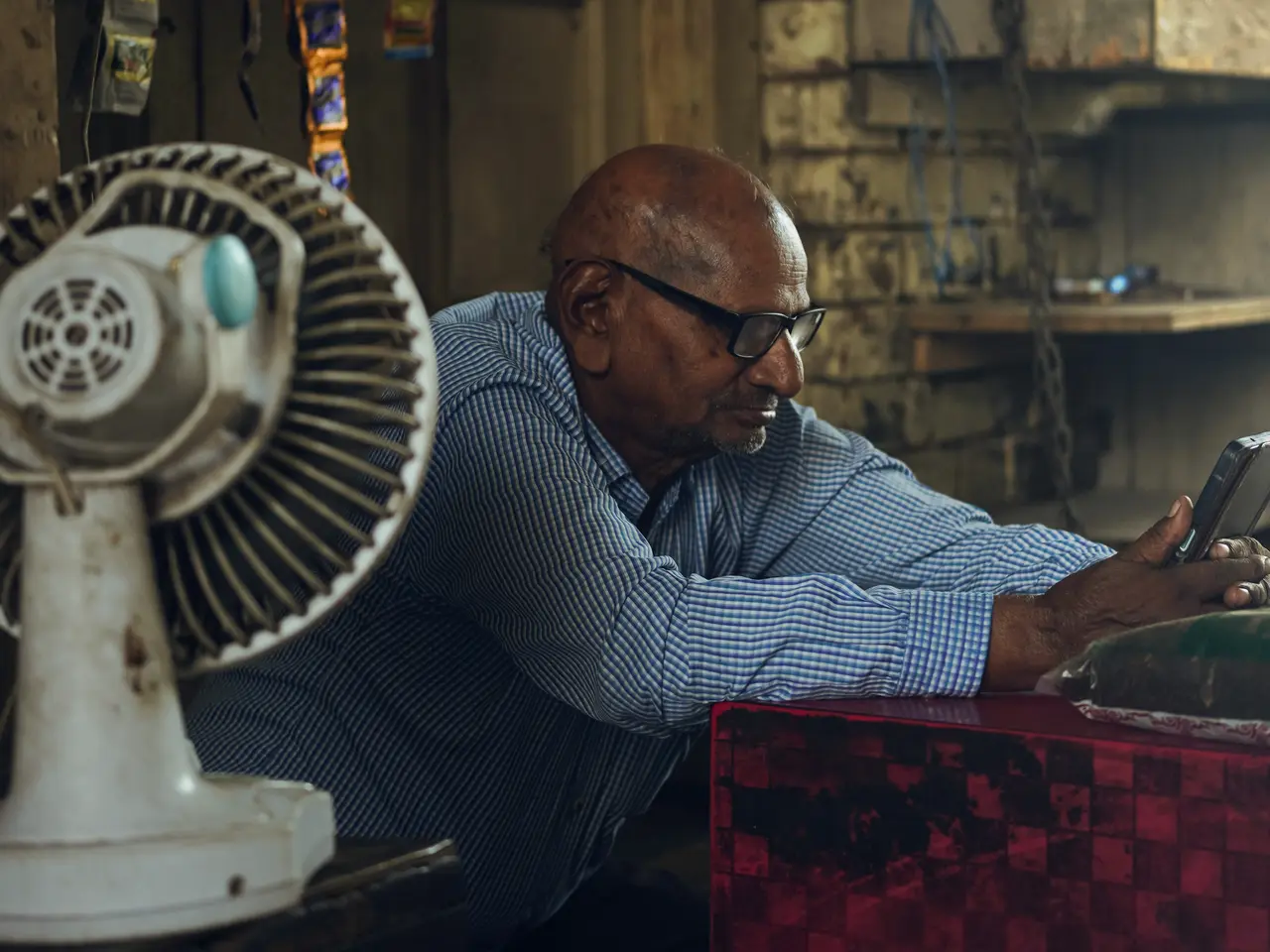An older shopkeeper in a checked shirt leans on his counter in a dim shop in Moga, Punjab, reading his phone beside a white table fan.