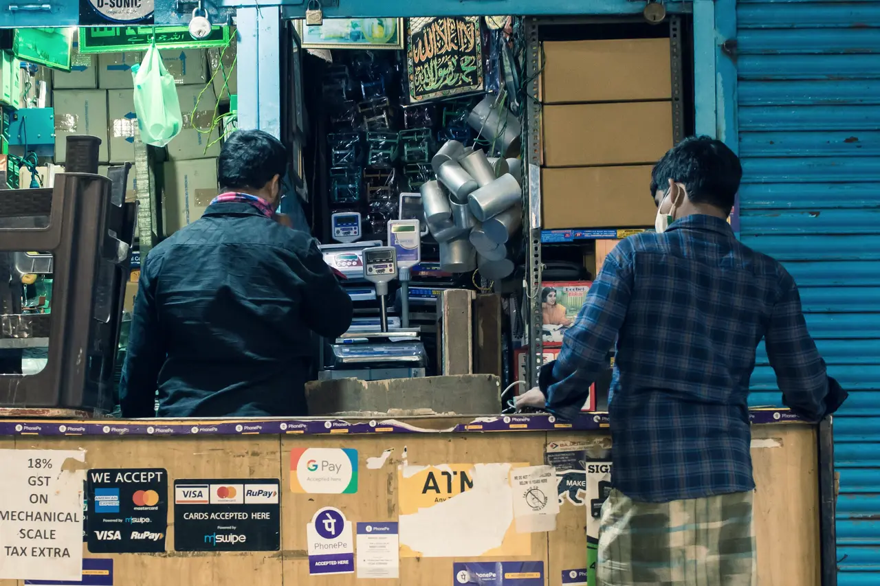 Two customers lean on a plywood shop counter in Bengaluru, between blue rolling shutters. The front of the counter is covered in payment stickers and a handwritten GST notice.