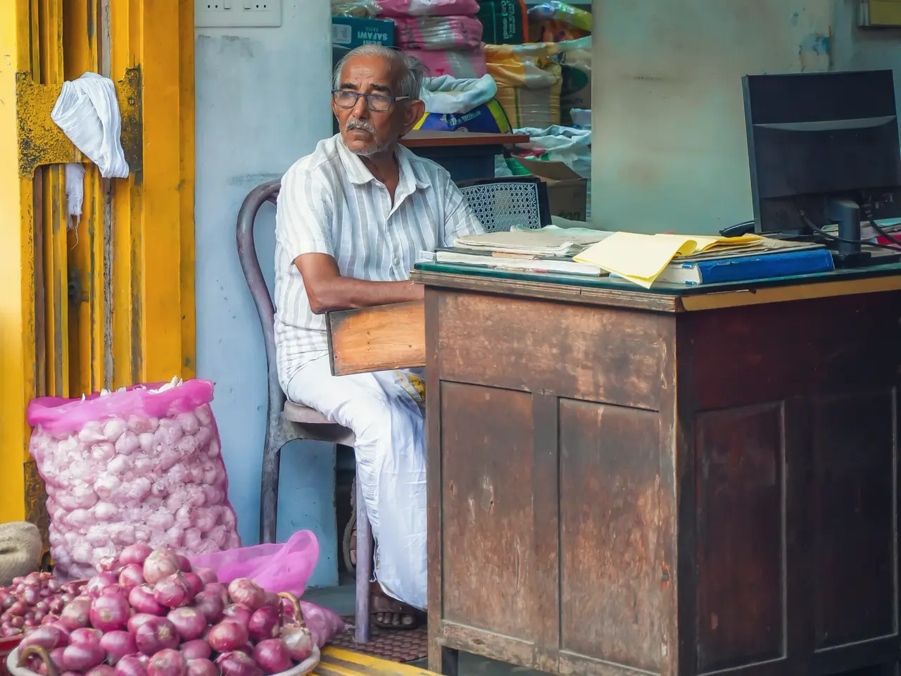 An older shopkeeper dressed in white sits at a worn wooden desk with files and a computer monitor, inside a small grocery shop in Thiruvananthapuram, with a basket of onions at the door.