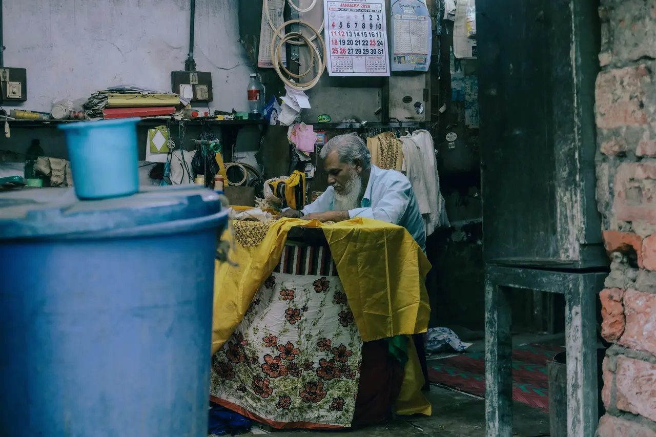 An elderly tailor with a white beard bends over his sewing machine in a cluttered workroom in Kolkata, with a blue plastic drum and bucket in front and a wall calendar above.