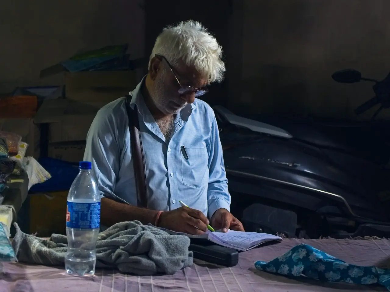 An older man with white hair and glasses writes in a small notebook at a table in a dim room, a water bottle and a folded cloth beside him.